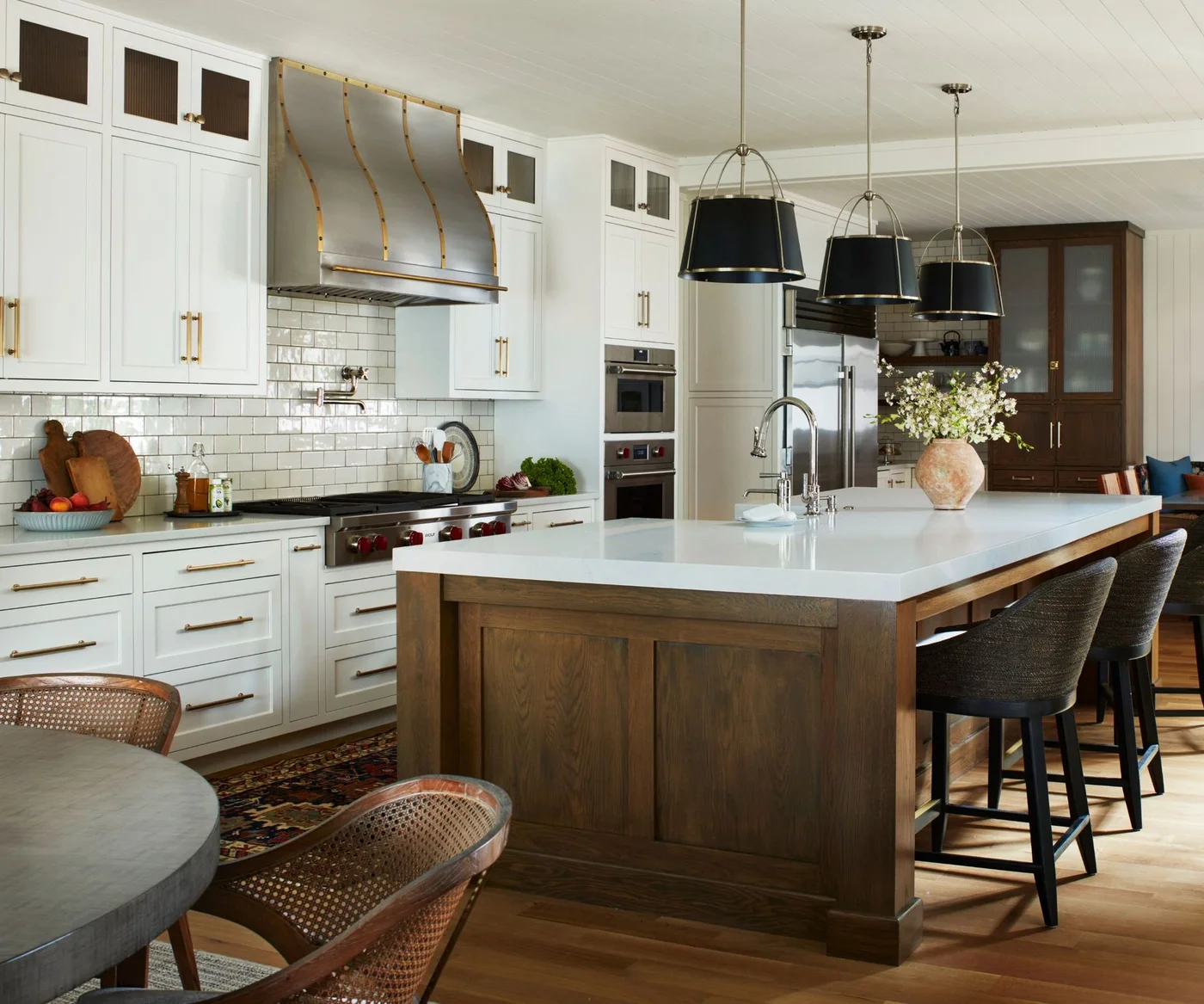 A white kitchen has a curved metal hood with warm-colored trim, three dark pendants, a wood island and a separate dark glazed cabinet.