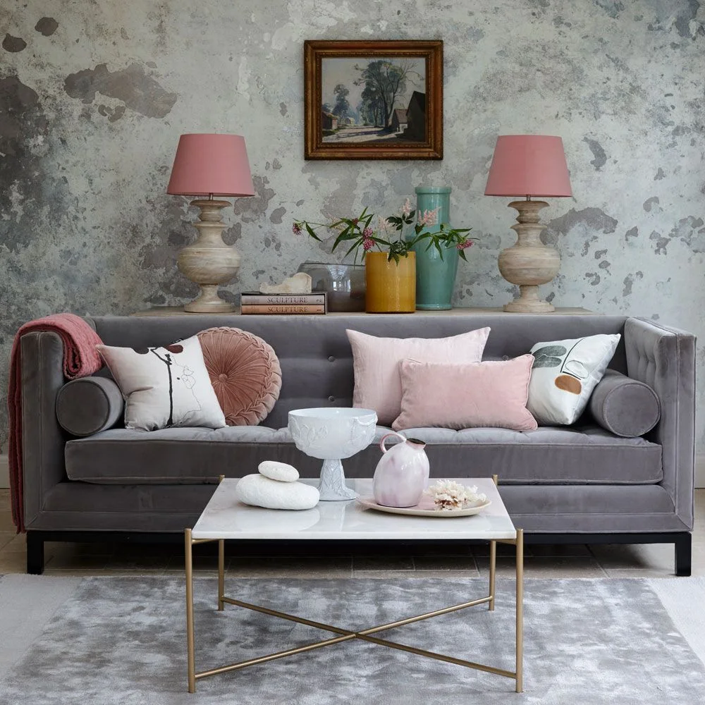 A dark gray sofa with rounded bolster cushions sits against a mottled gray wall between pink-shaded lamps behind a pale coffee table