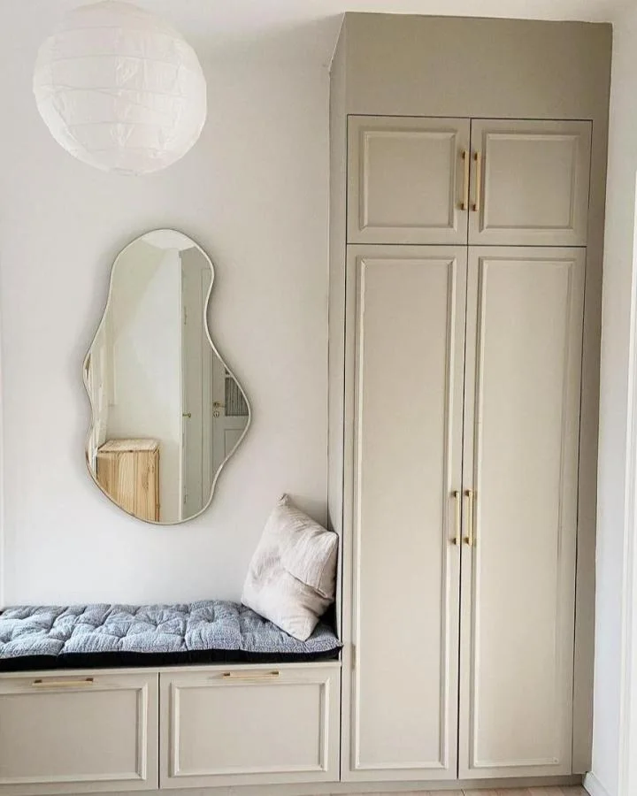 Pale paneled mudroom cabinet with gold-toned handles beside a gray-cushioned low unit and an irregularly shaped mirror.