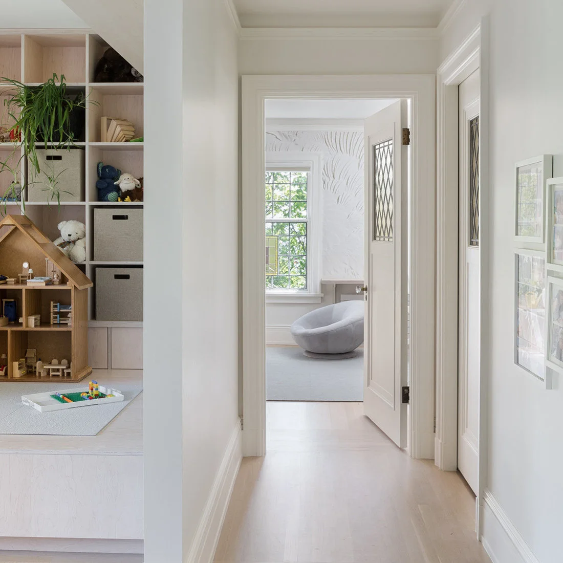A pale hallway passes a play area with tall cube storage, fabric bins, a wood dollhouse and a shallow toy tray on a floor mat.