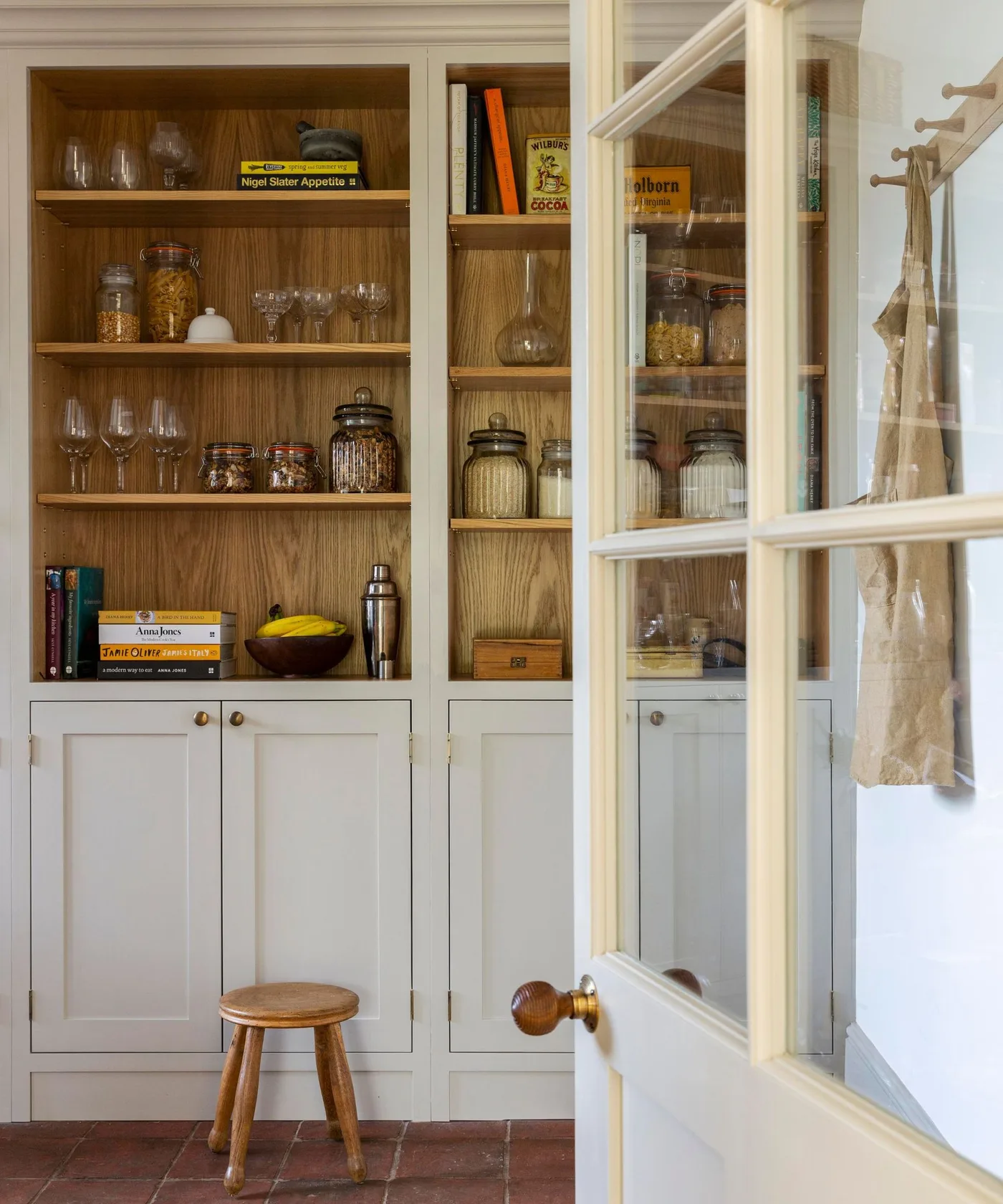 Country pantry with warm wood shelf interiors, pale lower cupboards, jar storage and glazed door