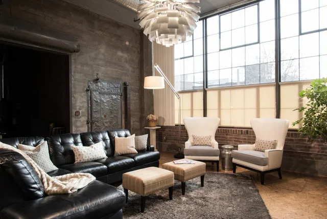 Black leather sofas face pale wingback chairs beside a broad window, exposed brick and two small stools