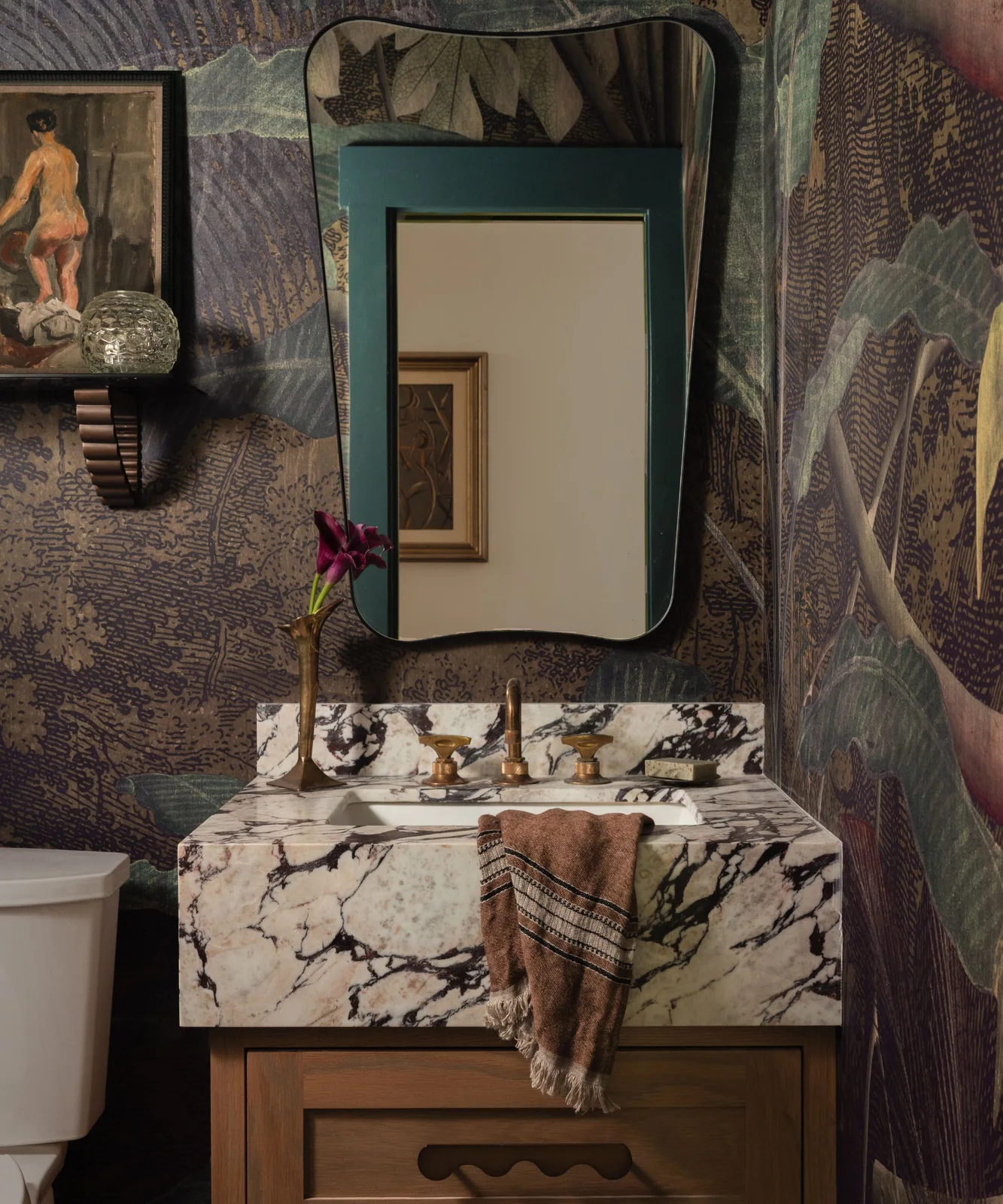A small powder room has dark botanical mural walls, an irregular black mirror, richly veined pale basin counter and a wood drawer cabinet.