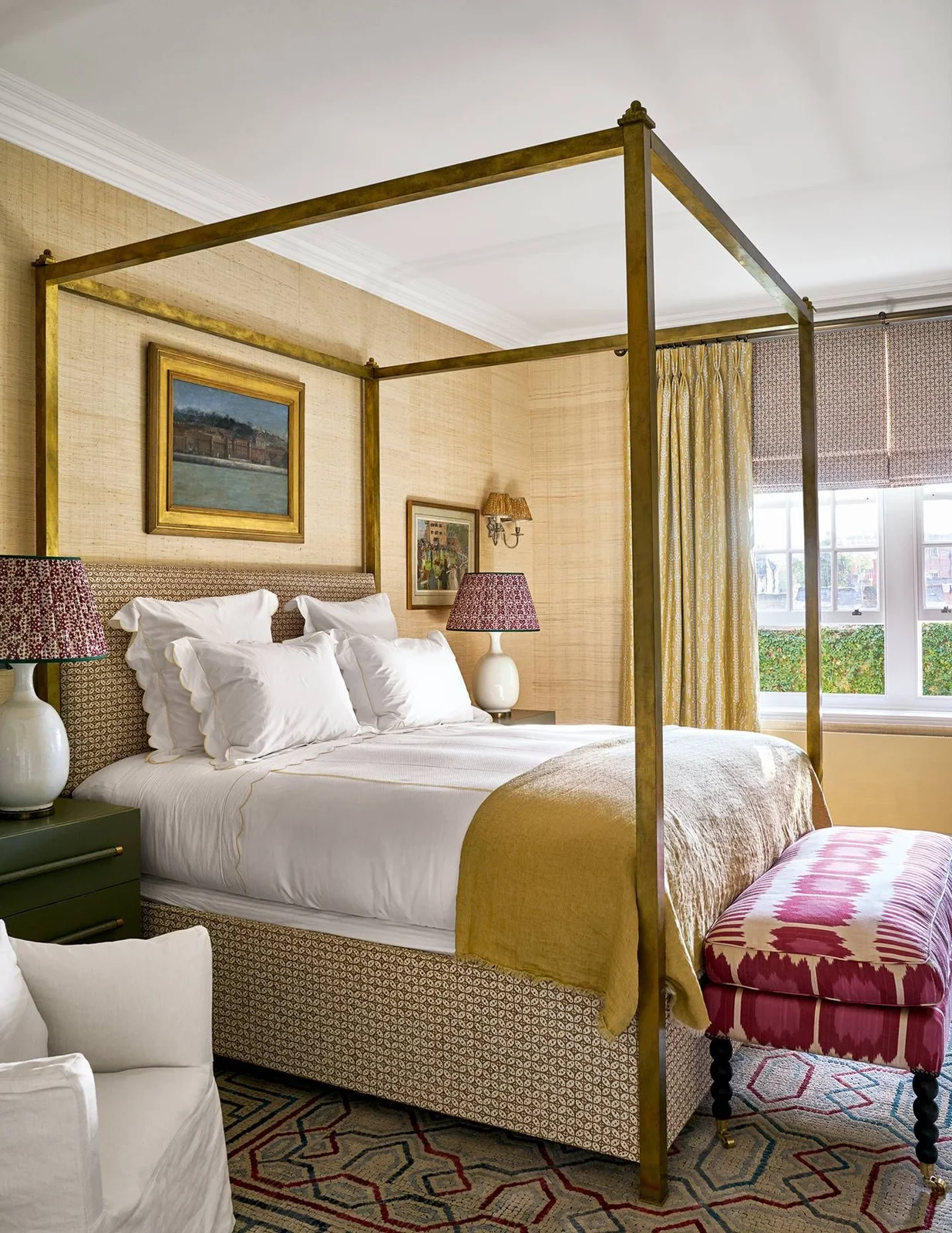 A gold-colored four-poster bed stands against a textured golden wall beside green bedside cabinets, patterned lampshades and a pink ottoman.