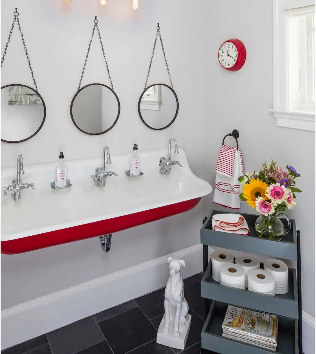 A dark towel ring holds a red-striped towel at the end of a long white trough sink with three faucets and circular mirrors.