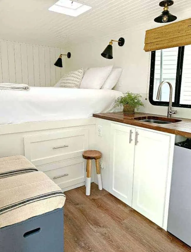 Compact white camper with drawers below a raised bed, wall reading lights and a small sink counter alongside.