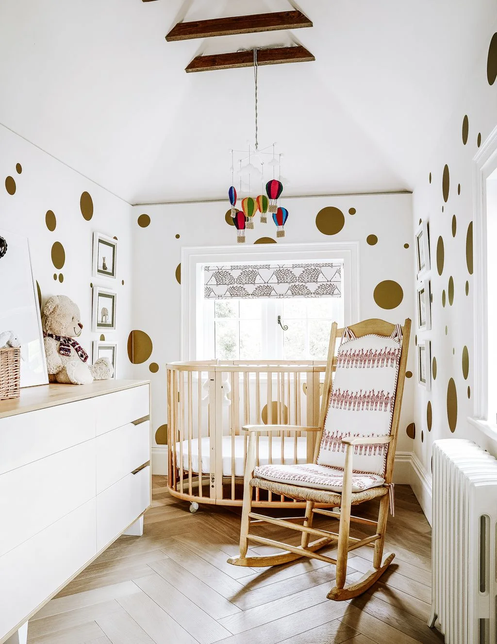 Large gold-colored wall dots surround a wooden nursery crib and patterned rocking chair beneath a colorful balloon-shaped hanging decoration