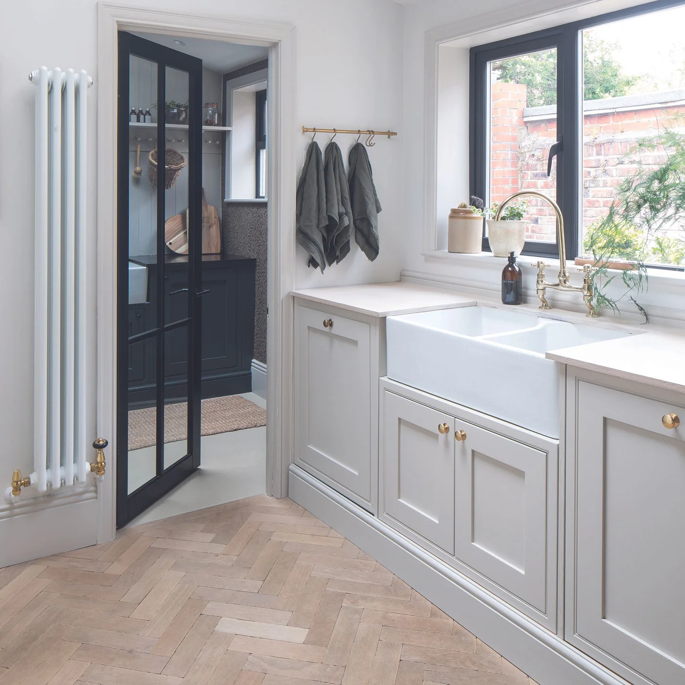 Pale gray drawers and paneled doors sit around a white apron-front sink beside dark-framed glazing and a light patterned wooden floor