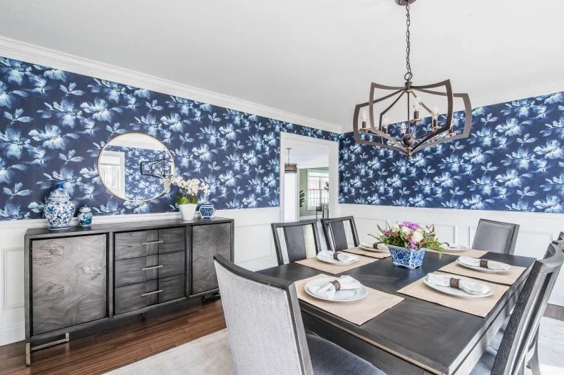 Pale upholstered dining chairs with dark rectangular frames around a dark table, against blue floral wallpaper above white paneling and a gray sideboard.