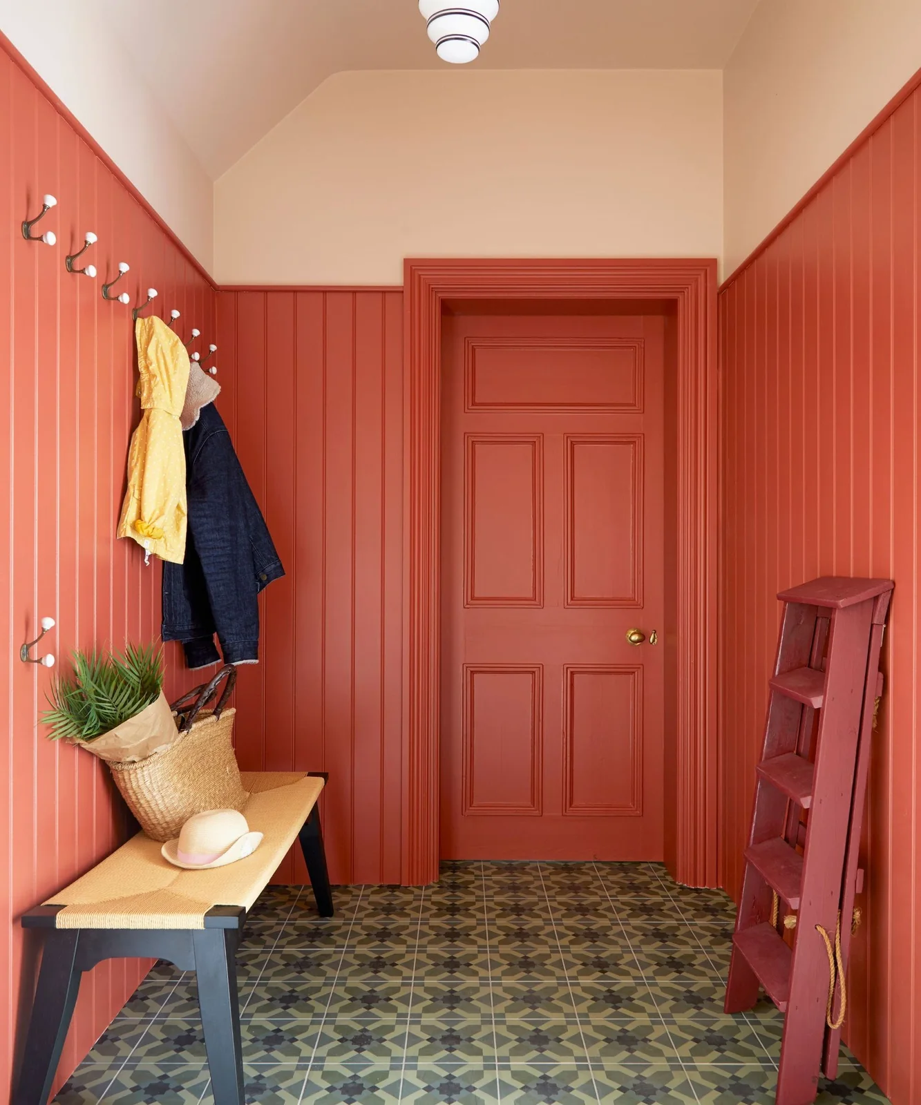 A plain wood entry bench stands beside coral vertical boards and two rows of hooks, with a coral paneled door, patterned floor and a small ladder