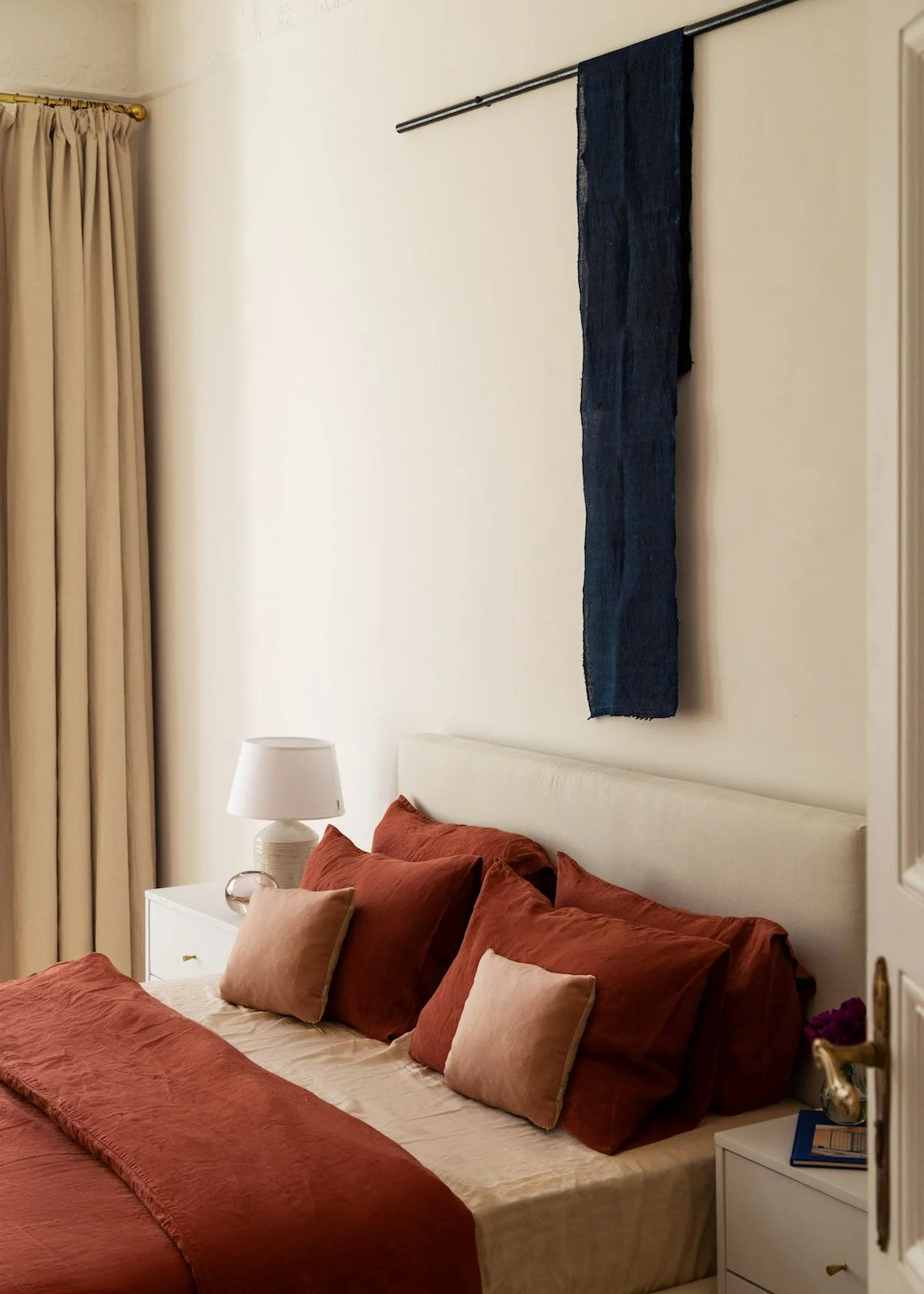 Cream bedroom with rust cushions and a rust bed runner, pale pink pillows and a narrow dark wall hanging