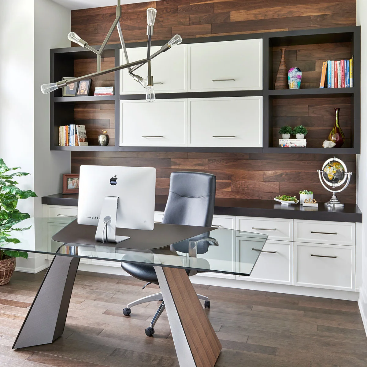 Glass-top desk on angled supports with a monitor and black chair before white cabinets and wood wall panels