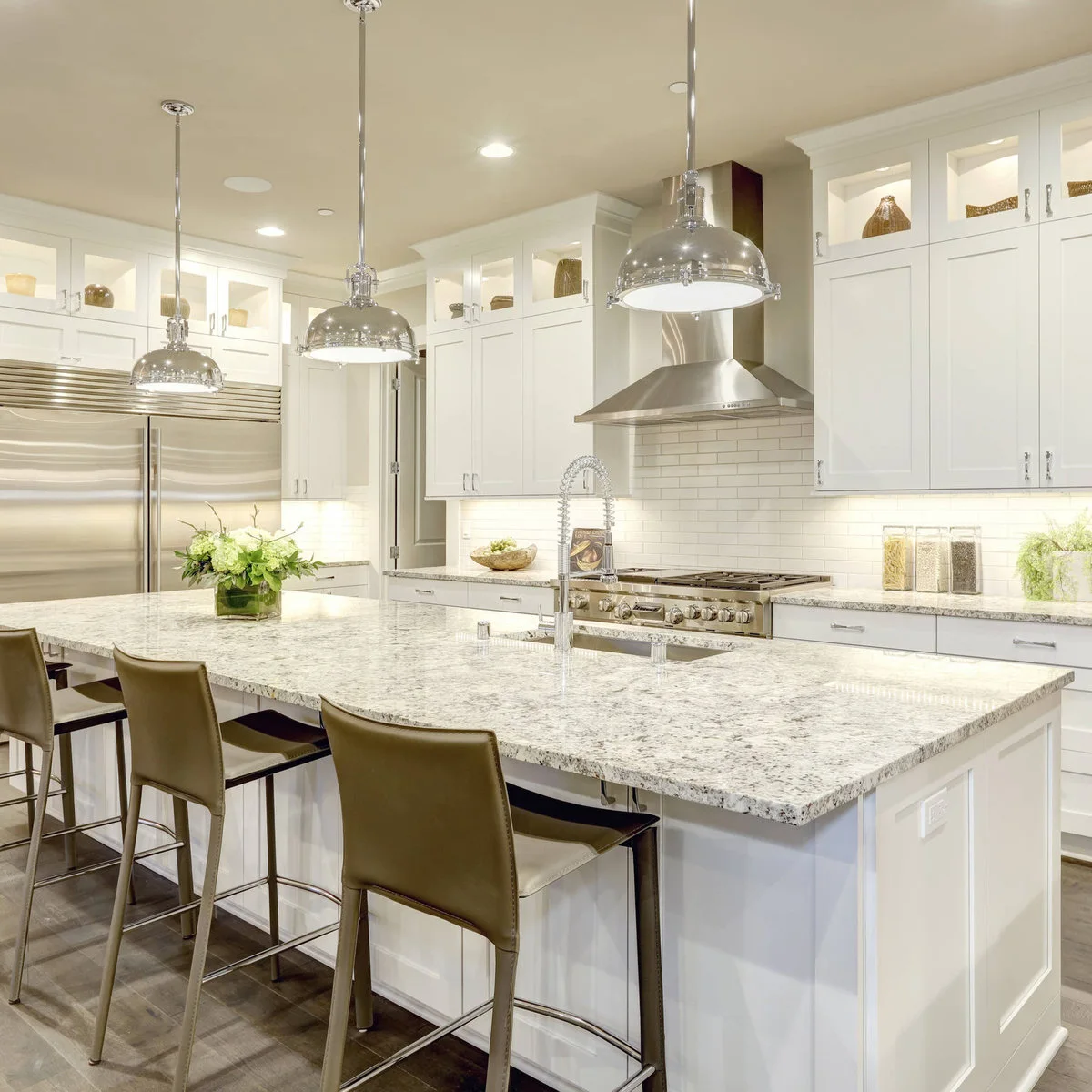 Shiny metal dome pendants hang above a pale island beneath white cupboards with lit glass-front display sections and illuminated counters.