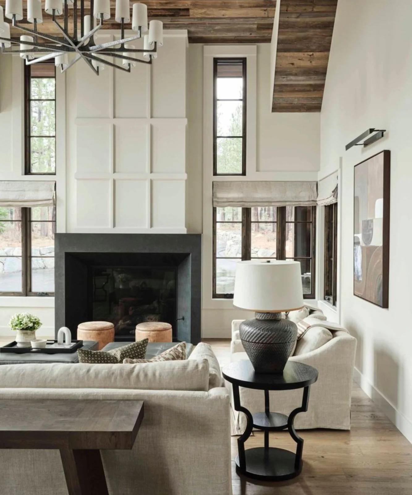 A pale sectional and dark round side table sit beneath a timber-clad ceiling beside tall windows and a dark fireplace.