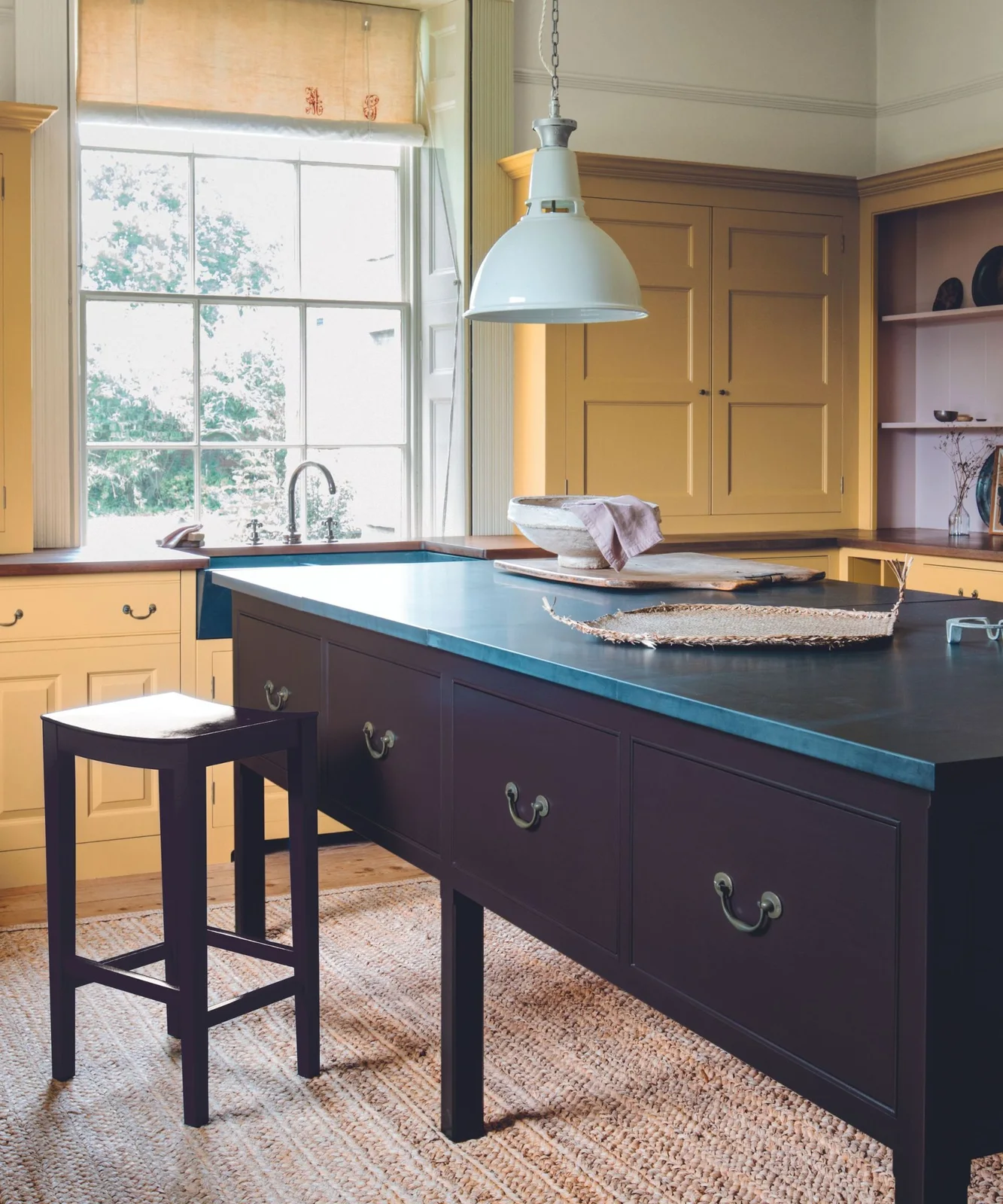 Mustard yellow cupboards surround a deep plum freestanding island with dark counters, a simple stool and a large sink window.