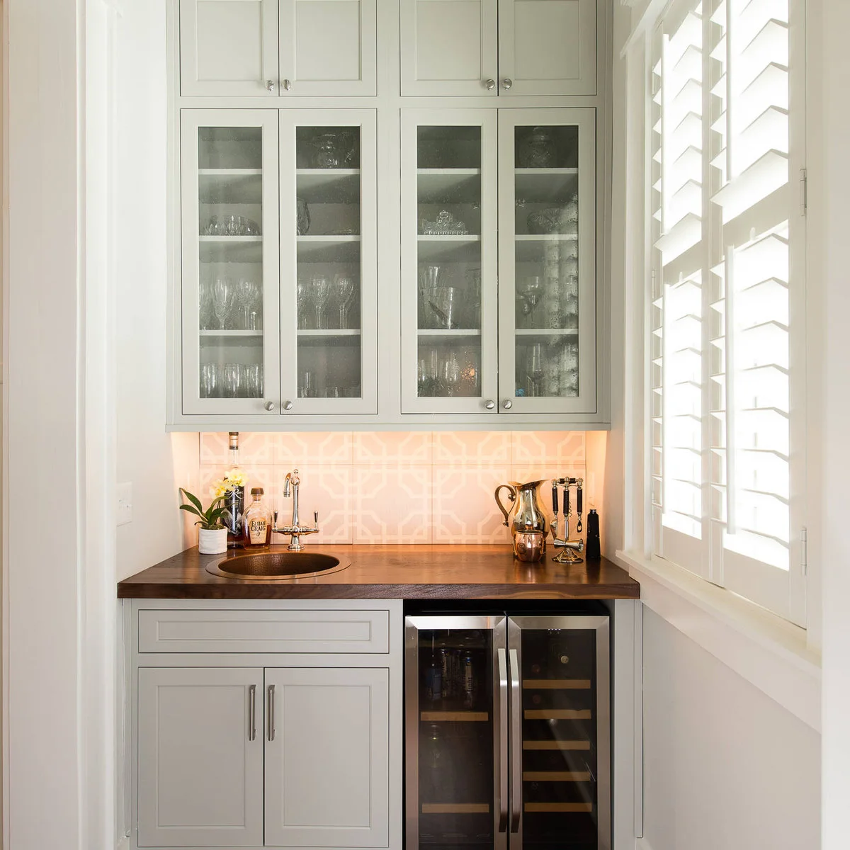 Pale gray mini bar with round sink, dark wood counter, patterned pale backsplash and glass-front upper cupboards