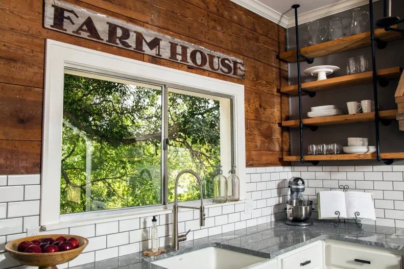 Country sink beneath large window with wood-board wall above white rectangular tile, open dark-framed shelves and a wood sign