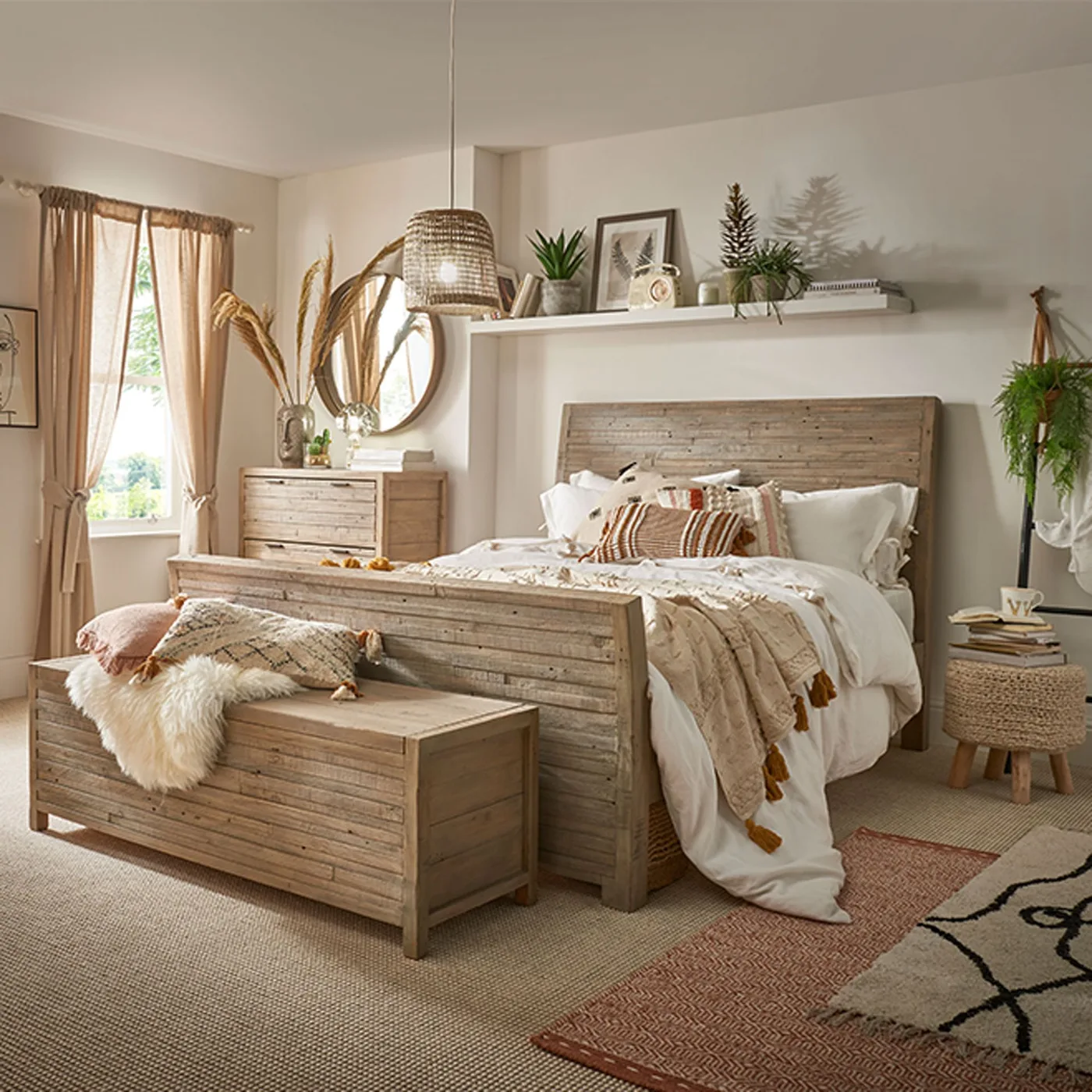 A wood bed, matching chest and blanket box stand beneath a long shelf with plants beside pale bedding, woven shades and a small patterned rug