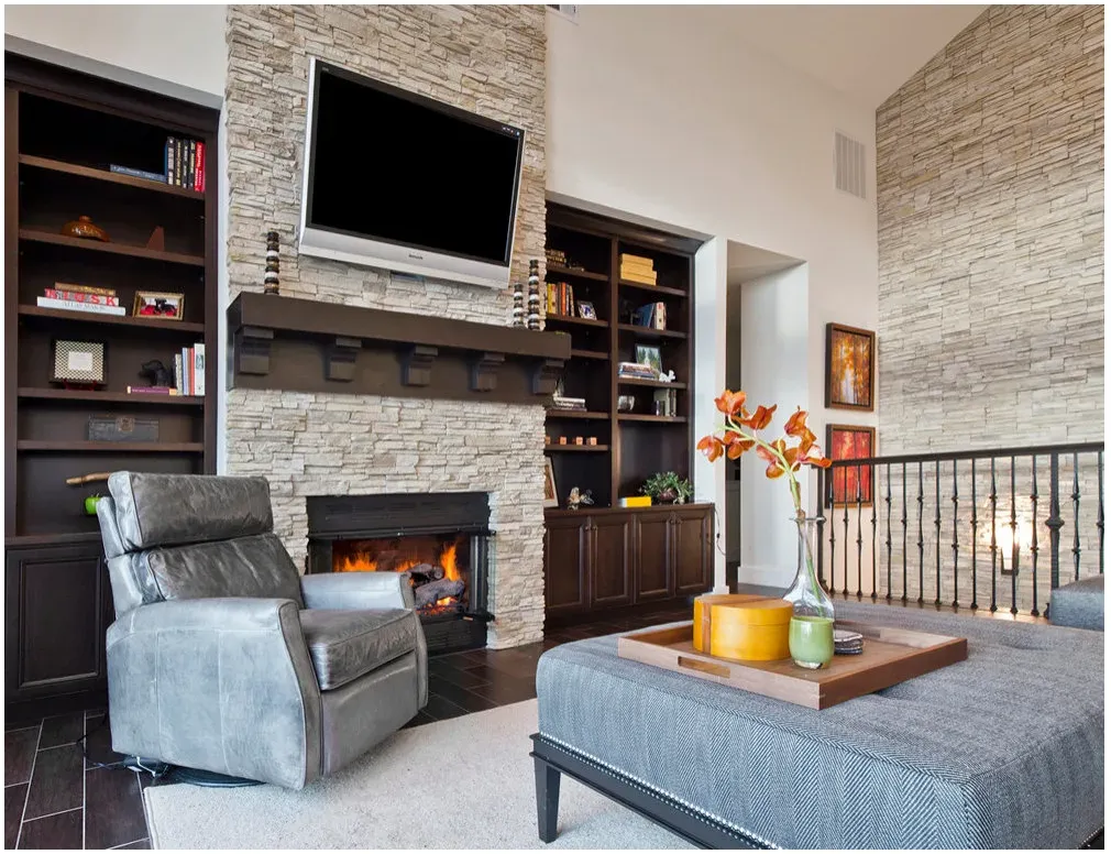 A television above a dark mantel on a tall textured fireplace wall, with dark built-in shelves and a gray chair.