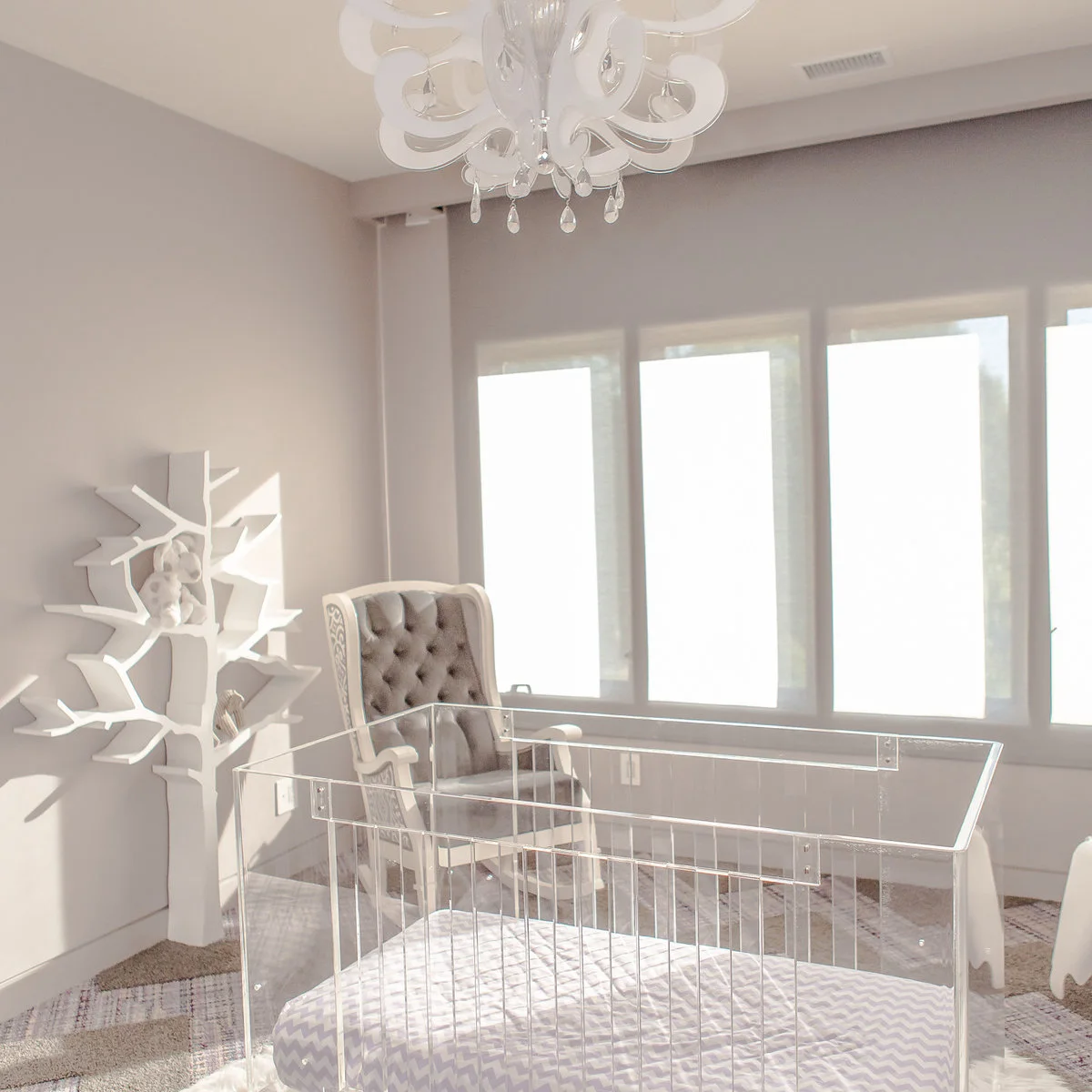Pale nursery with a transparent crib, grey tufted chair and white tree-shaped wall shelves