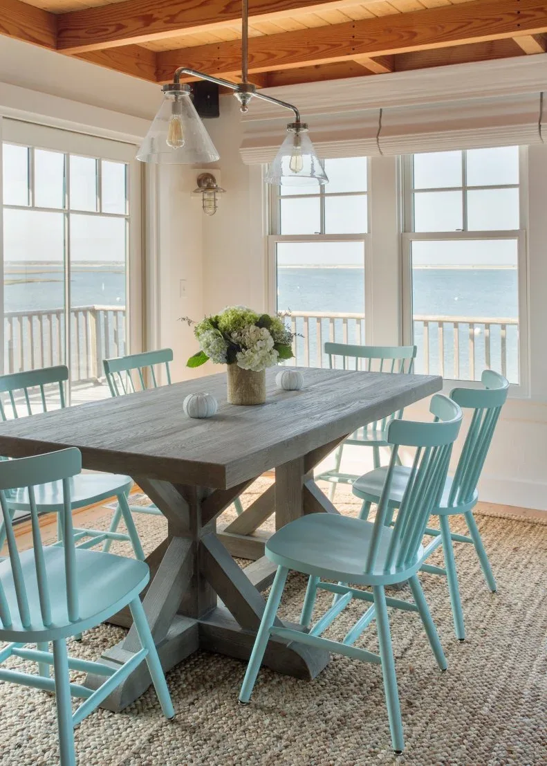 Pale blue spindle chairs surround a gray trestle dining table beneath an exposed timber ceiling and clear pendant lights.