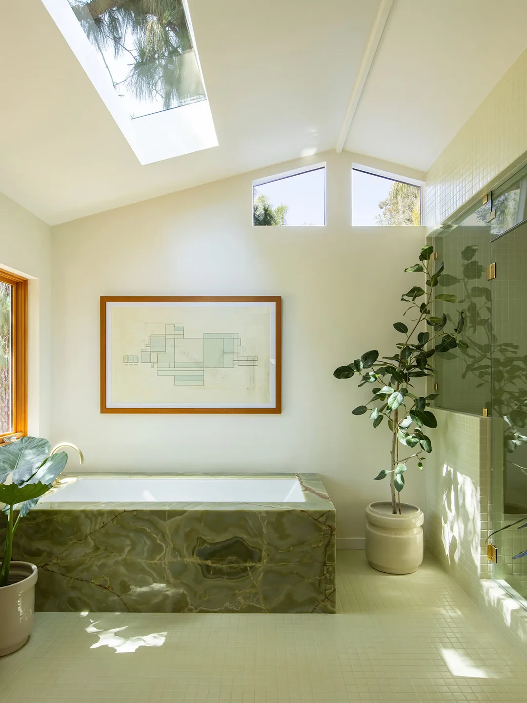 A rectangular bath with a green veined surround sits below high windows and a skylight beside a framed print, potted plants and glass shower