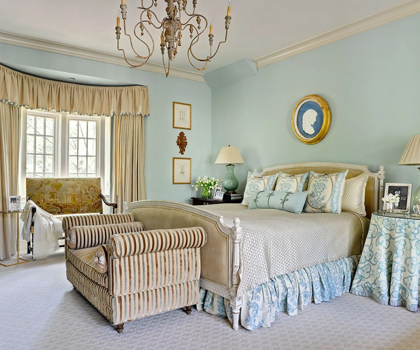 Country bedroom with pale blue walls, curved bed frame, patterned bed skirt, striped armchair, long cream curtains and a gold-colored ceiling light.