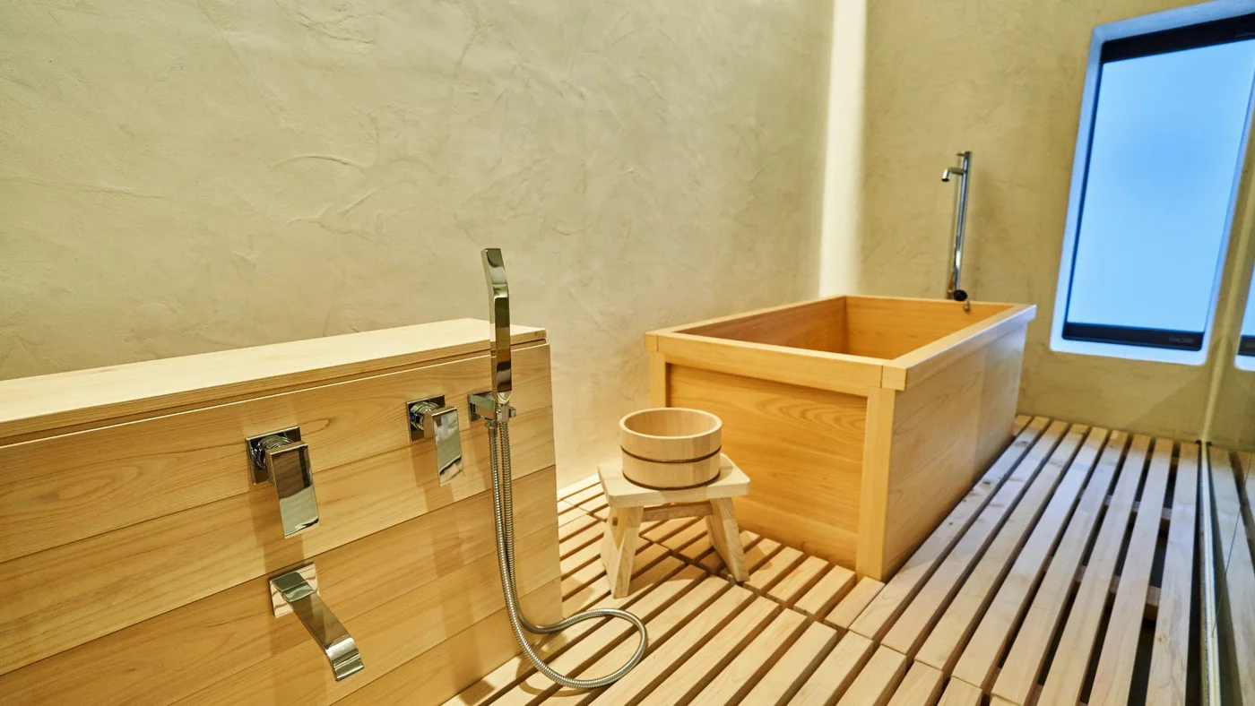 A rectangular wooden tub stands beside a low wooden stool and bucket, slatted floor and wall-mounted washing fittings beneath a small window