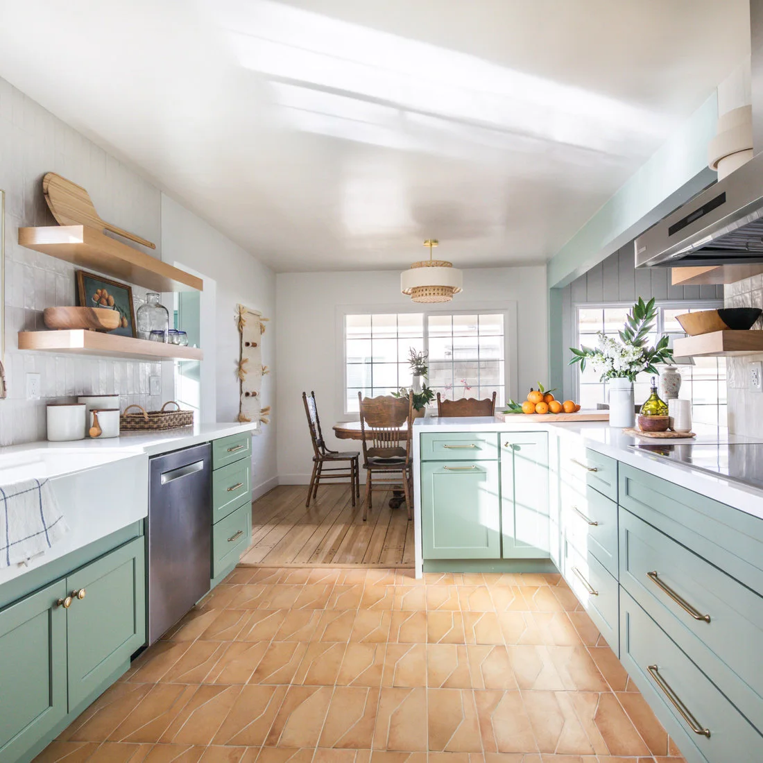 Peach terracotta rectangles cover a sunny kitchen floor between pale green cabinets and a dining doorway.
