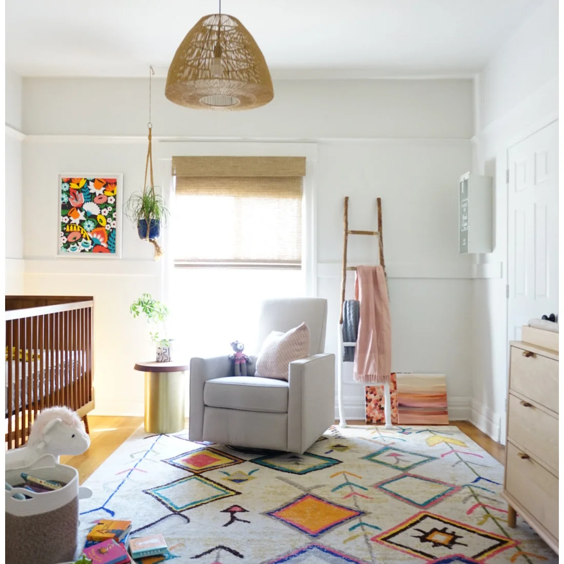 A gray armchair, small round side table and wood dresser sit on a diamond-patterned rug beneath a woven pendant in a white nursery.