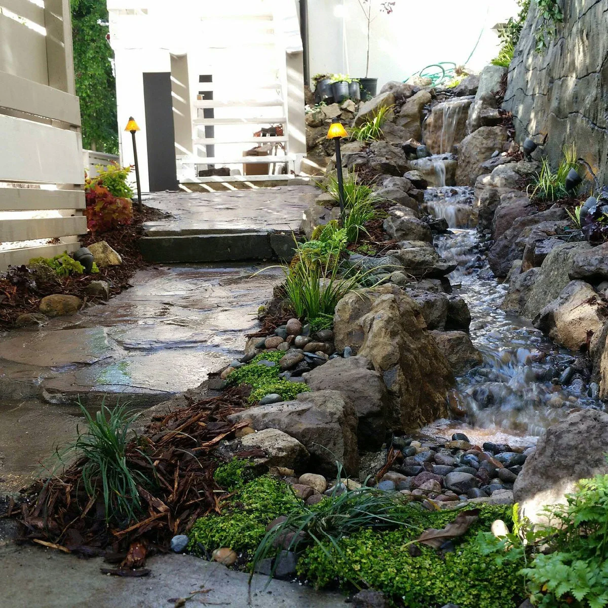 A narrow rock cascade beside a stone wall and paved side passage, with white fencing, small lights and young plants.