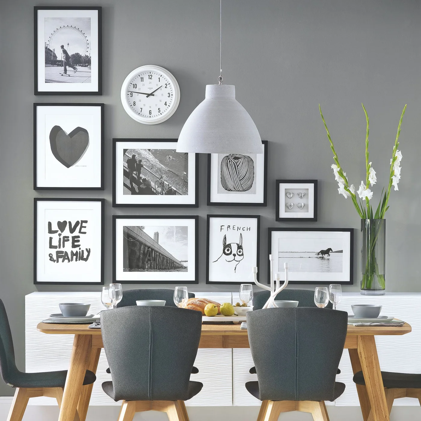 A low white textured sideboard holds a tall clear vase and white flowers beneath a black-framed gallery wall in a gray dining room.