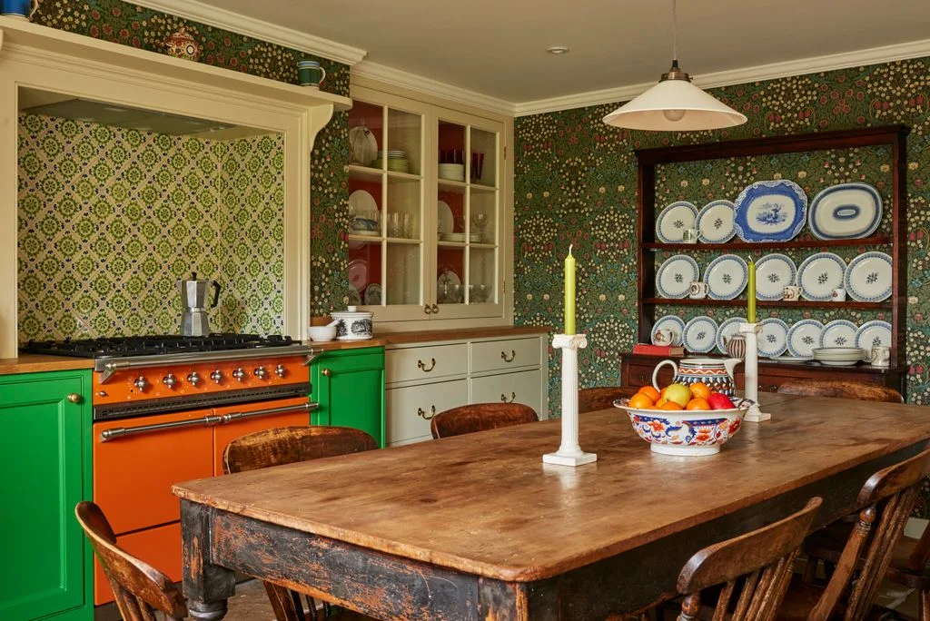 An orange range sits between bright green cabinets beneath floral wallpaper beside a plate-filled dresser and a wooden kitchen table