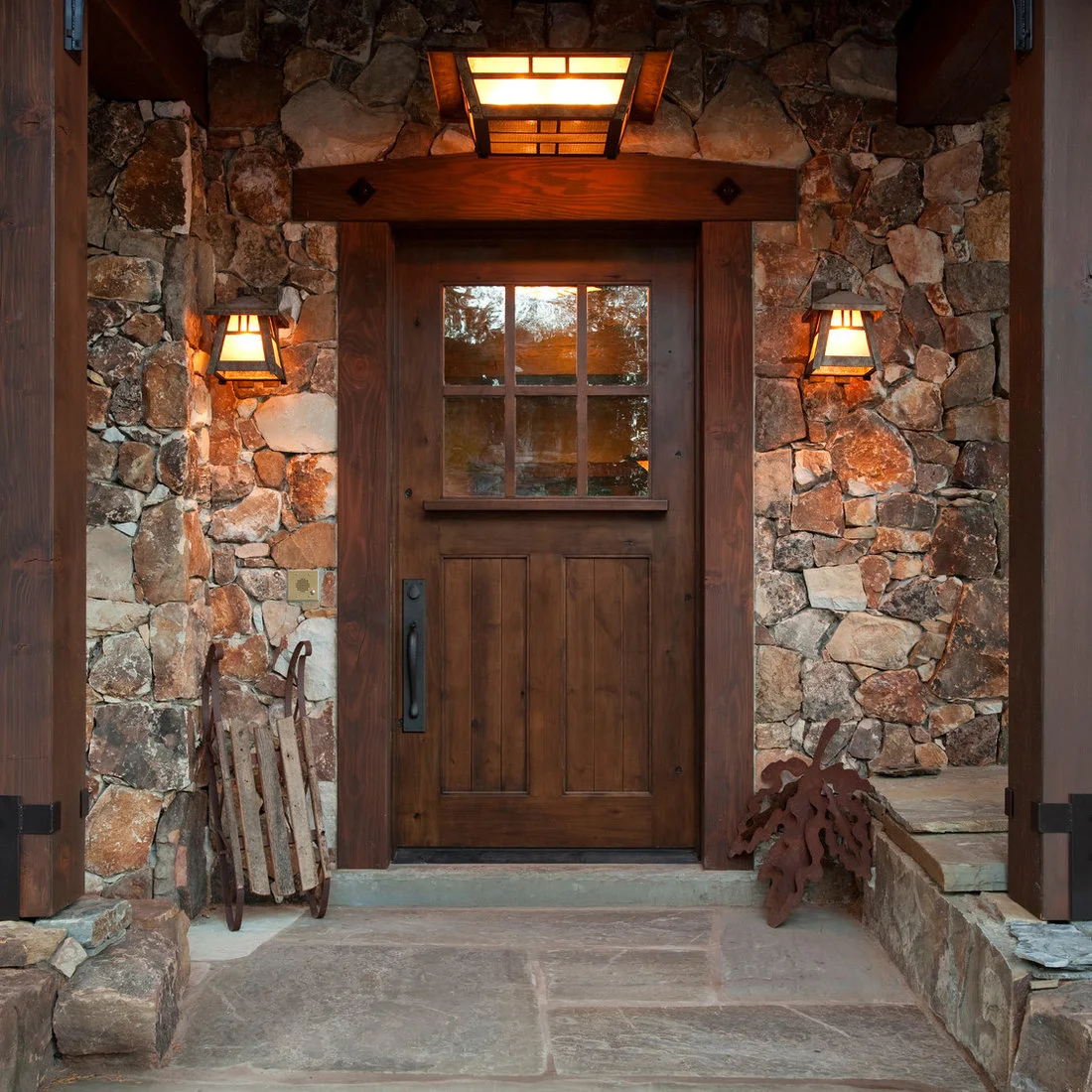 A timber door surrounded by rough stone beneath a large porch light and paired wall lamps