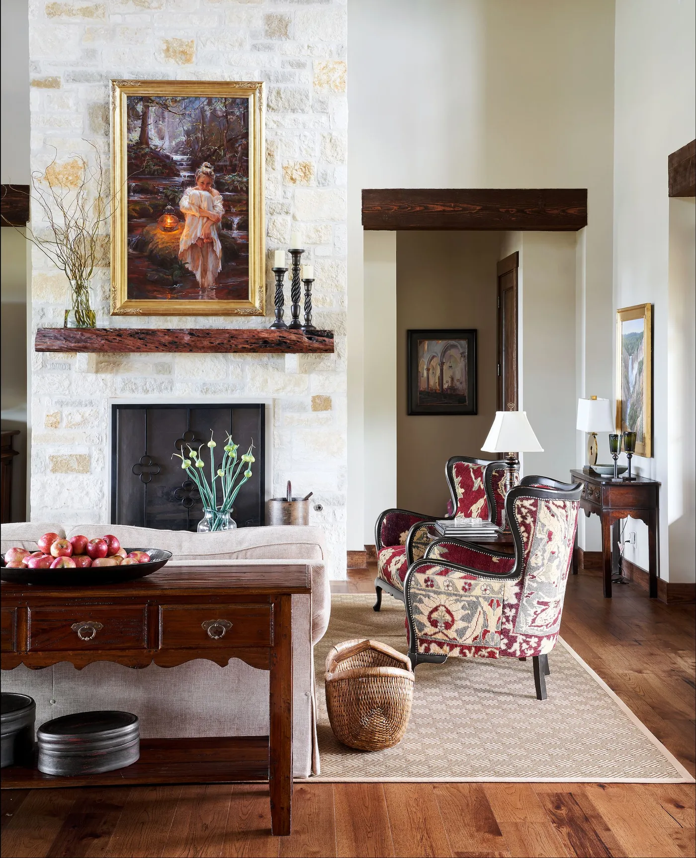 Tall pale stone chimney wall with narrow wooden mantel, framed picture, red-and-cream patterned chairs, table and woven basket.