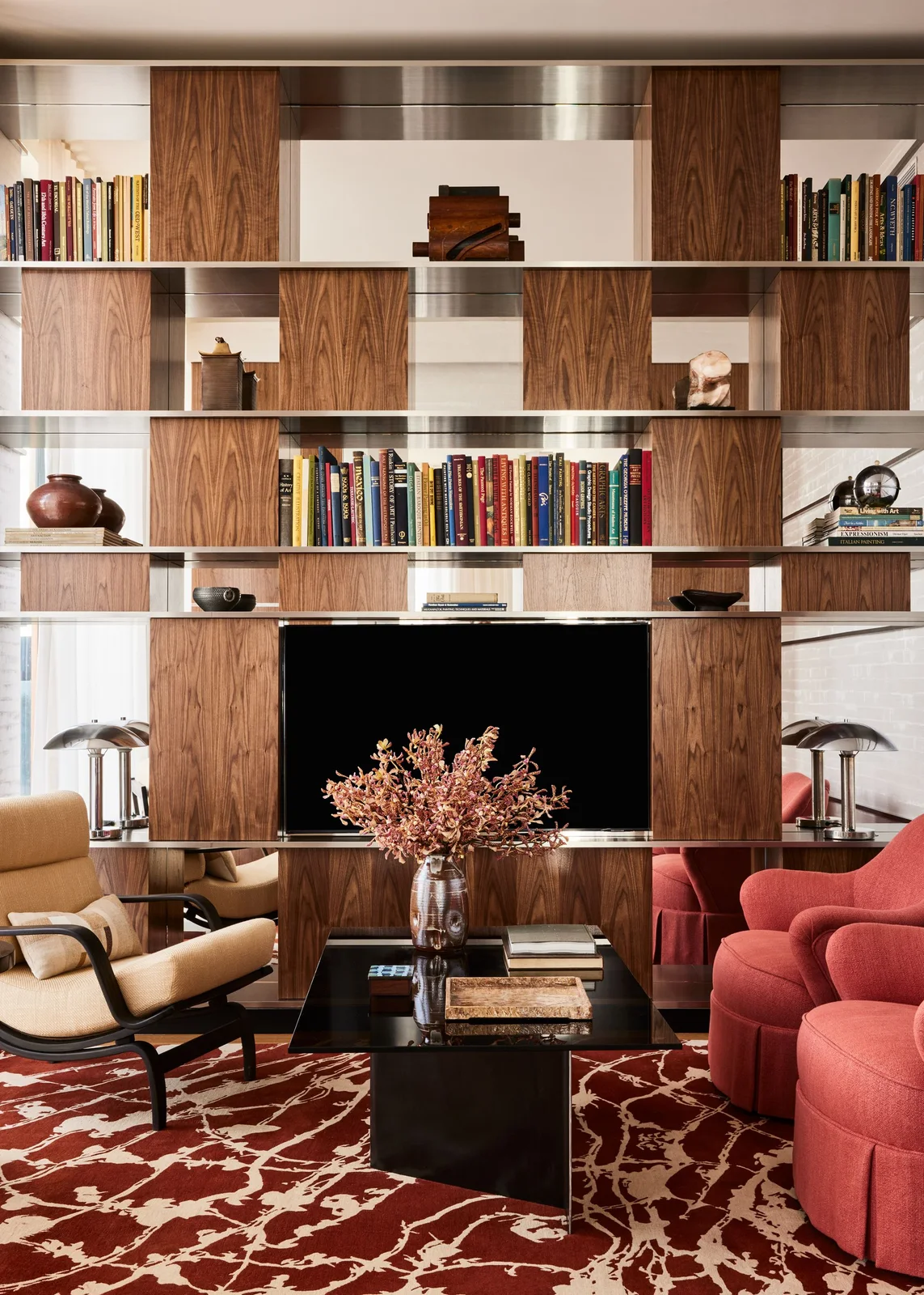 A television sits within a tall wooden and reflective shelving wall behind red chairs, a yellow chair and a dark table