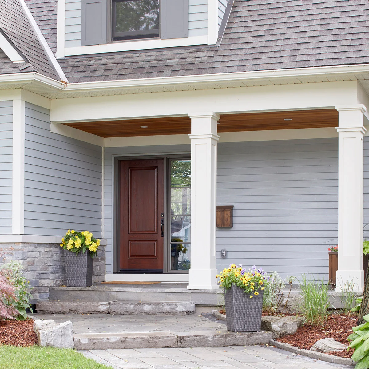 A gray-sided entry porch has white square posts, a brown door, a wood-toned ceiling and stone steps with yellow-flowered pots.