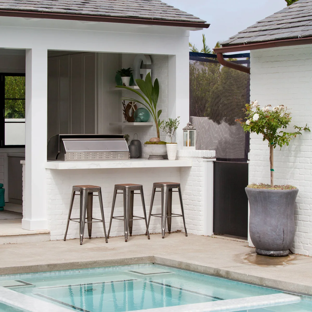 White poolside counter with three metal stools, open shelving, a closed grill and a large potted tree