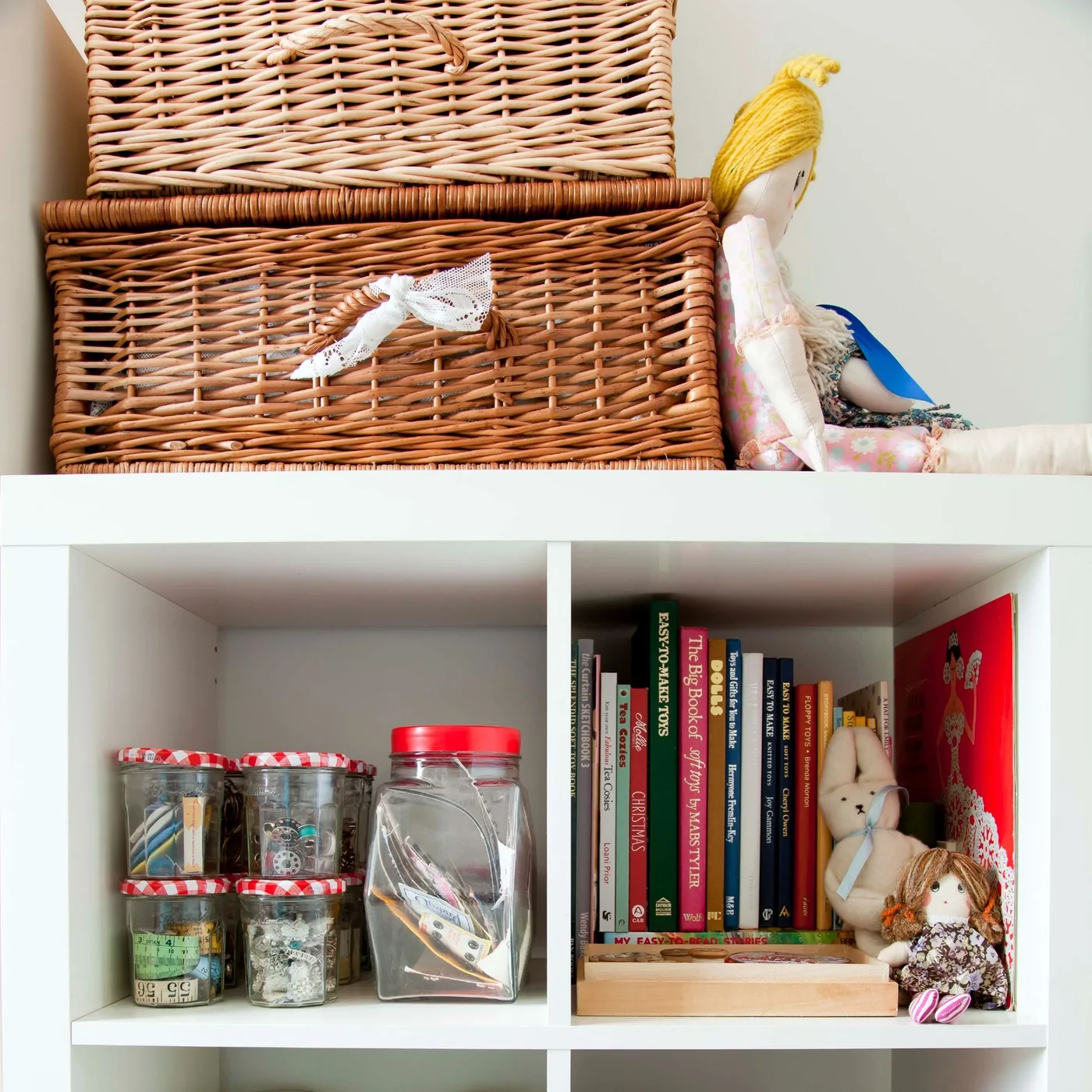White cube shelving with wicker baskets, dolls, books and jars containing sewing and craft supplies