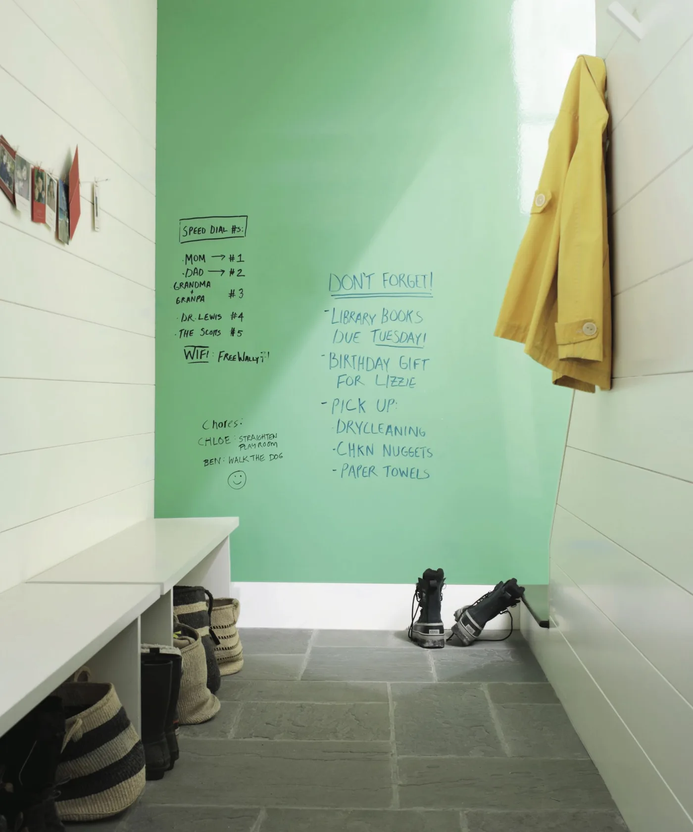 Narrow entry with green message wall, white bench and paneling, baskets underneath, hanging yellow coat and gray floor