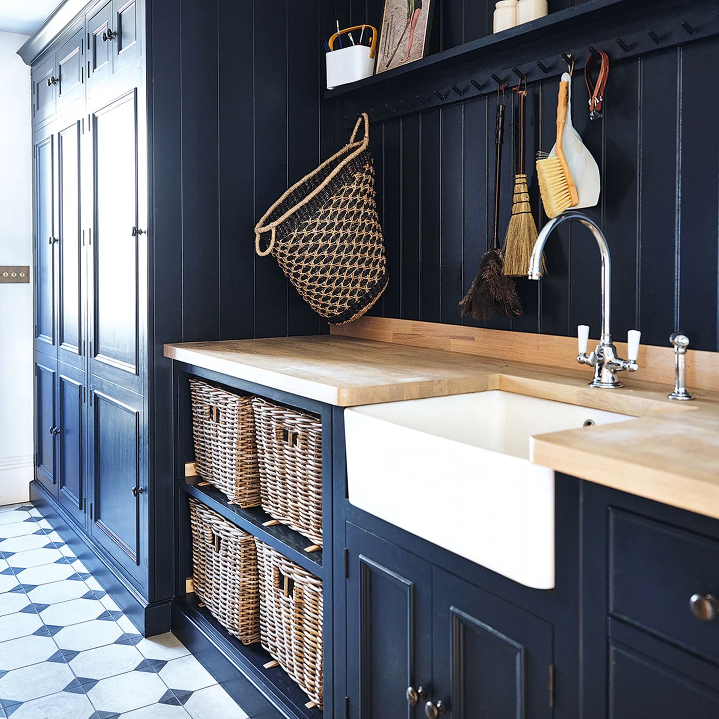 Navy paneled storage surrounds a white apron-front sink, wood-toned counter, woven baskets, hanging cleaning tools and patterned floor