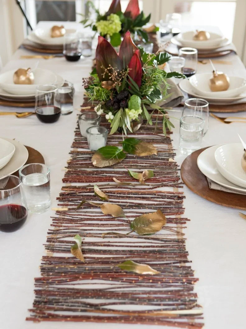 Green foliage and red-toned flowers sit above a long twig runner between pale place settings with warm metallic accents
