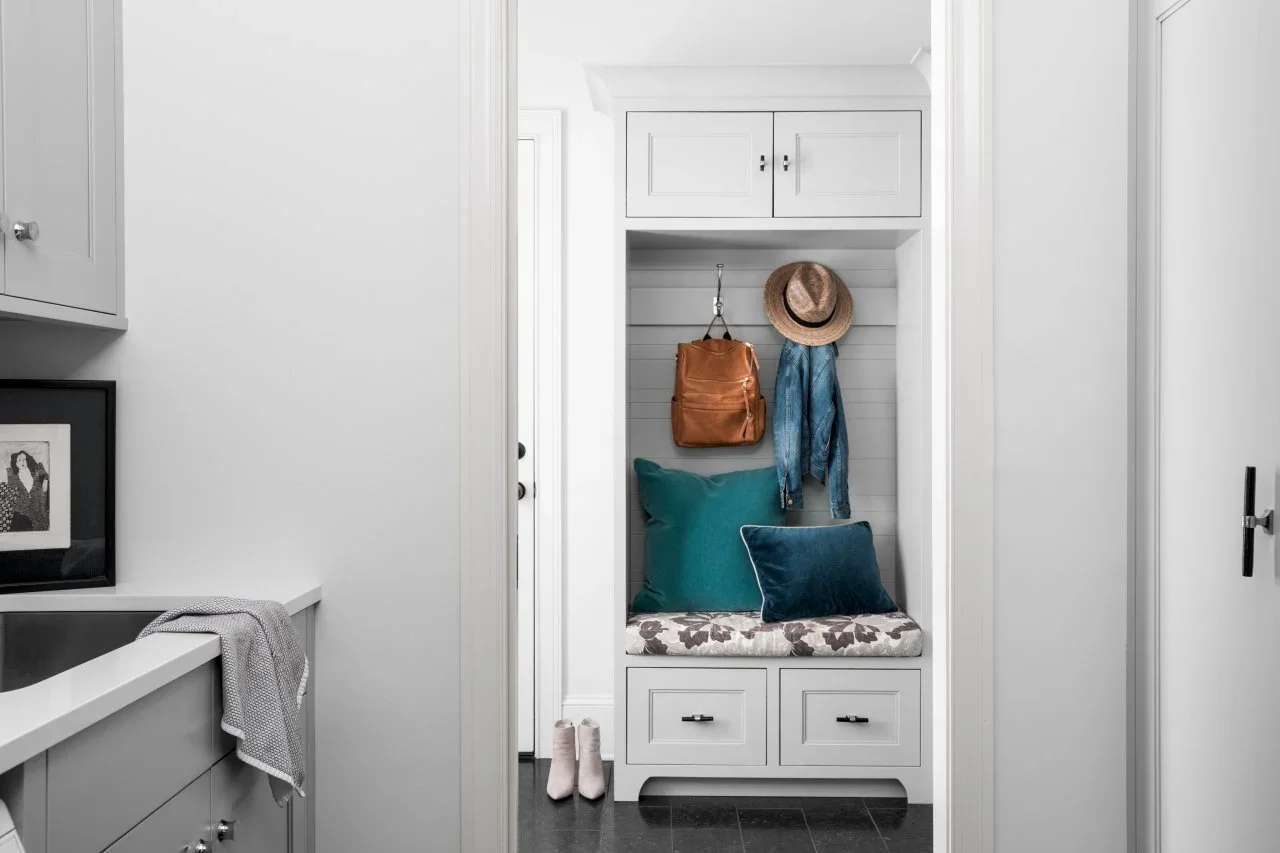 A view from a gray laundry counter looks through a pocket doorway toward a fitted entry bench with drawers, coat hooks and teal cushions.