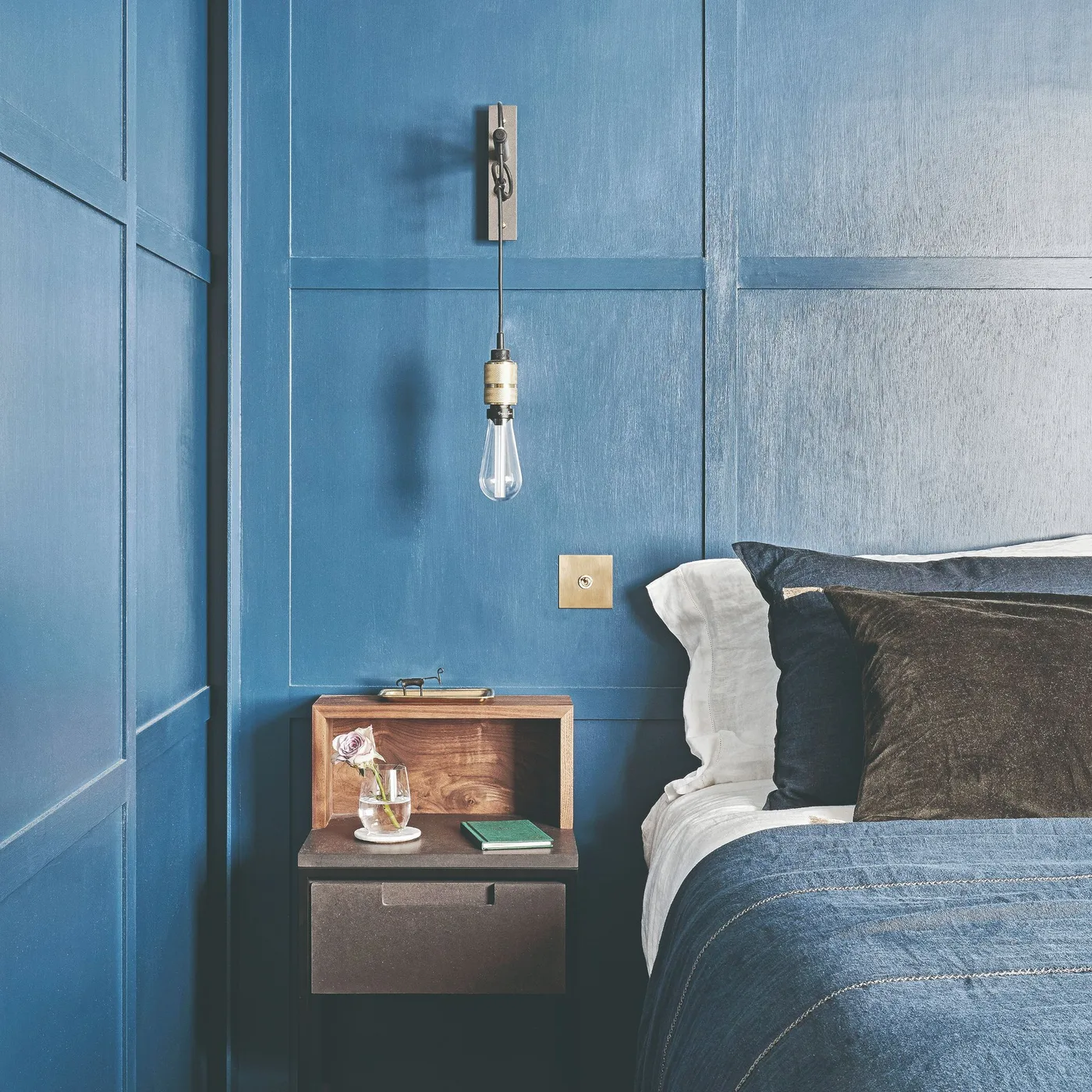Deep blue paneled bedroom wall with a floating wooden bedside table, an exposed-bulb pendant, and blue-gray bedding.