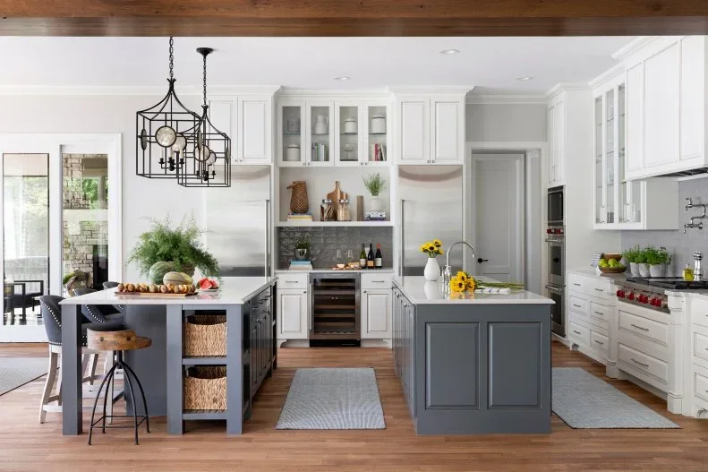 Two gray paneled islands stand within white kitchen cabinets and glass upper cupboards beneath open dark pendant frames beside warm flooring