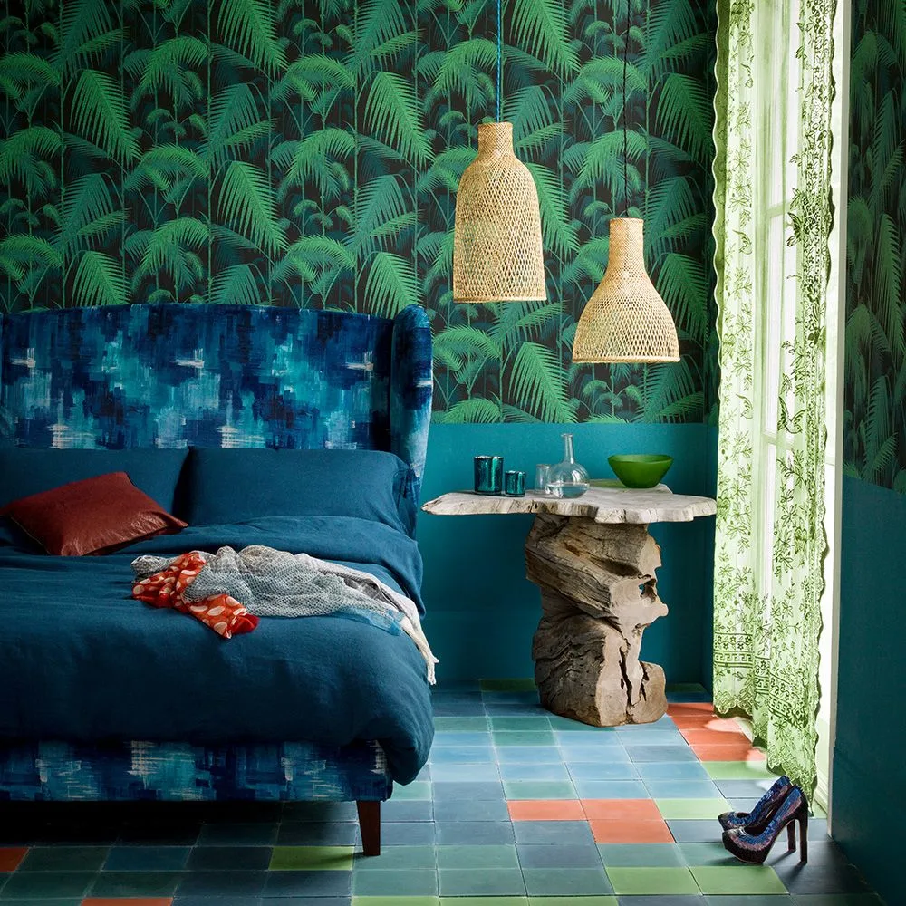 Dark palm wallpaper sits above a teal lower wall beside a blue upholstered bed, two woven pendant shades and a sculptural wood bedside table