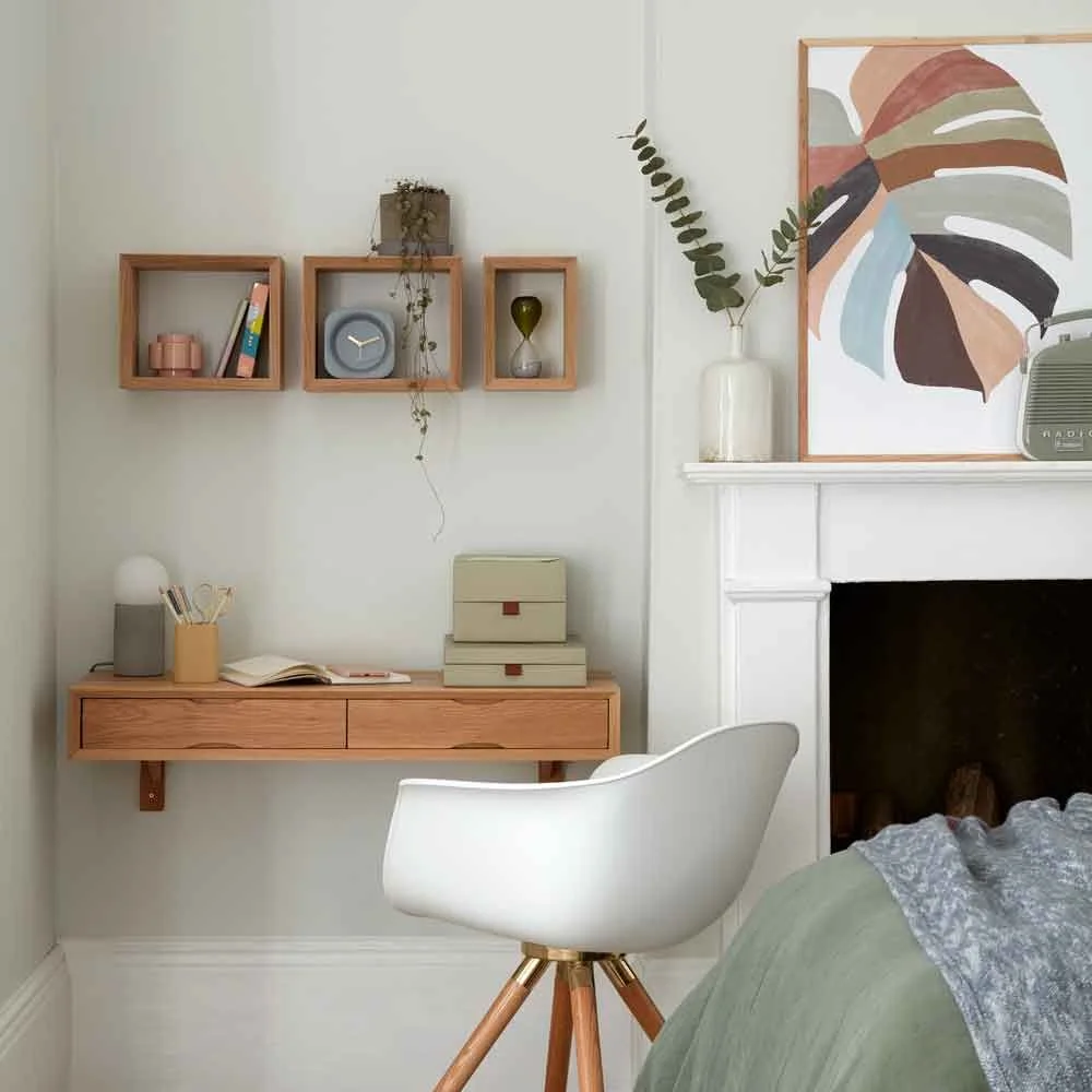 A small floating wood drawer desk and white wood-leg chair sit beside a white fireplace, with three square wood display shelves and muted artwork nearby.