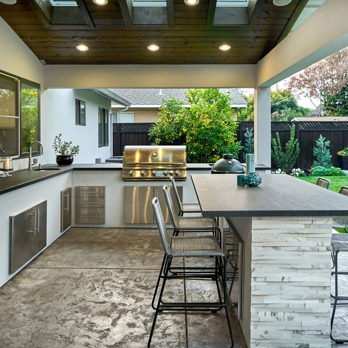 Rectangular gray outdoor bar island on a white textured base with slim woven stools and kitchen counters behind