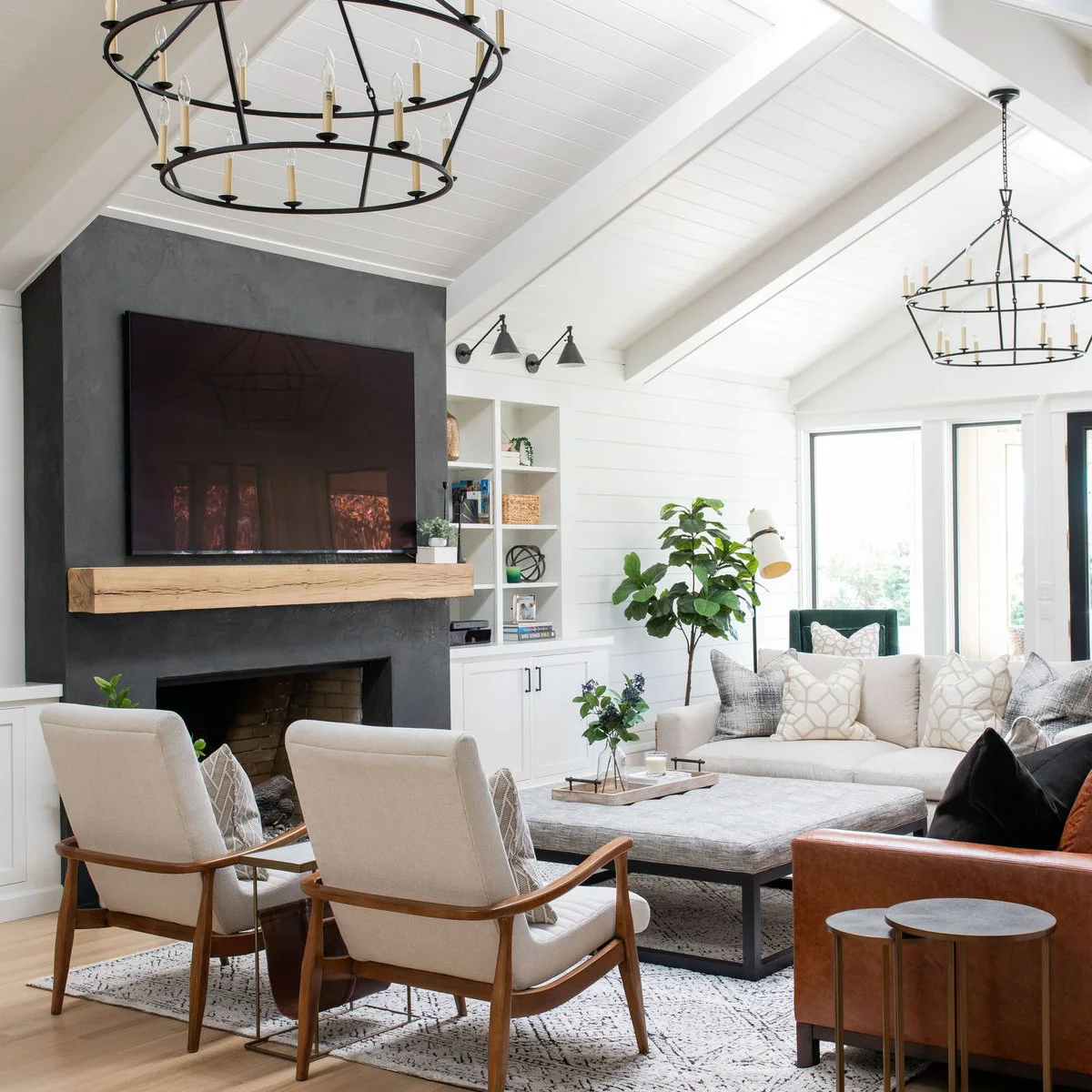 A large television above a pale wood mantel on a dark fireplace wall in a white room with open shelves and cream seating.