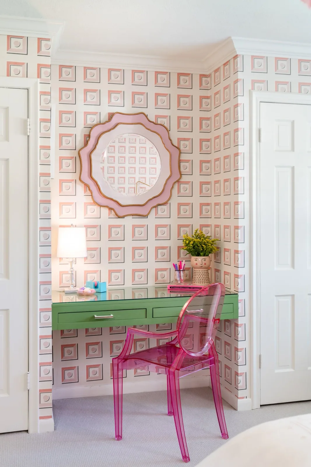Pink geometric wallpaper frames a green bedroom desk, transparent pink chair and scalloped mirror.
